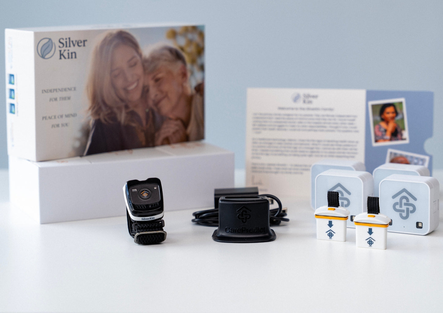 Silver Kin smartwatch care kit displayed on a white surface, including the retail box with a photo of an elderly woman, a black charging dock, a black smartwatch, two small white accessory cases, and printed instruction cards against a light blue background.