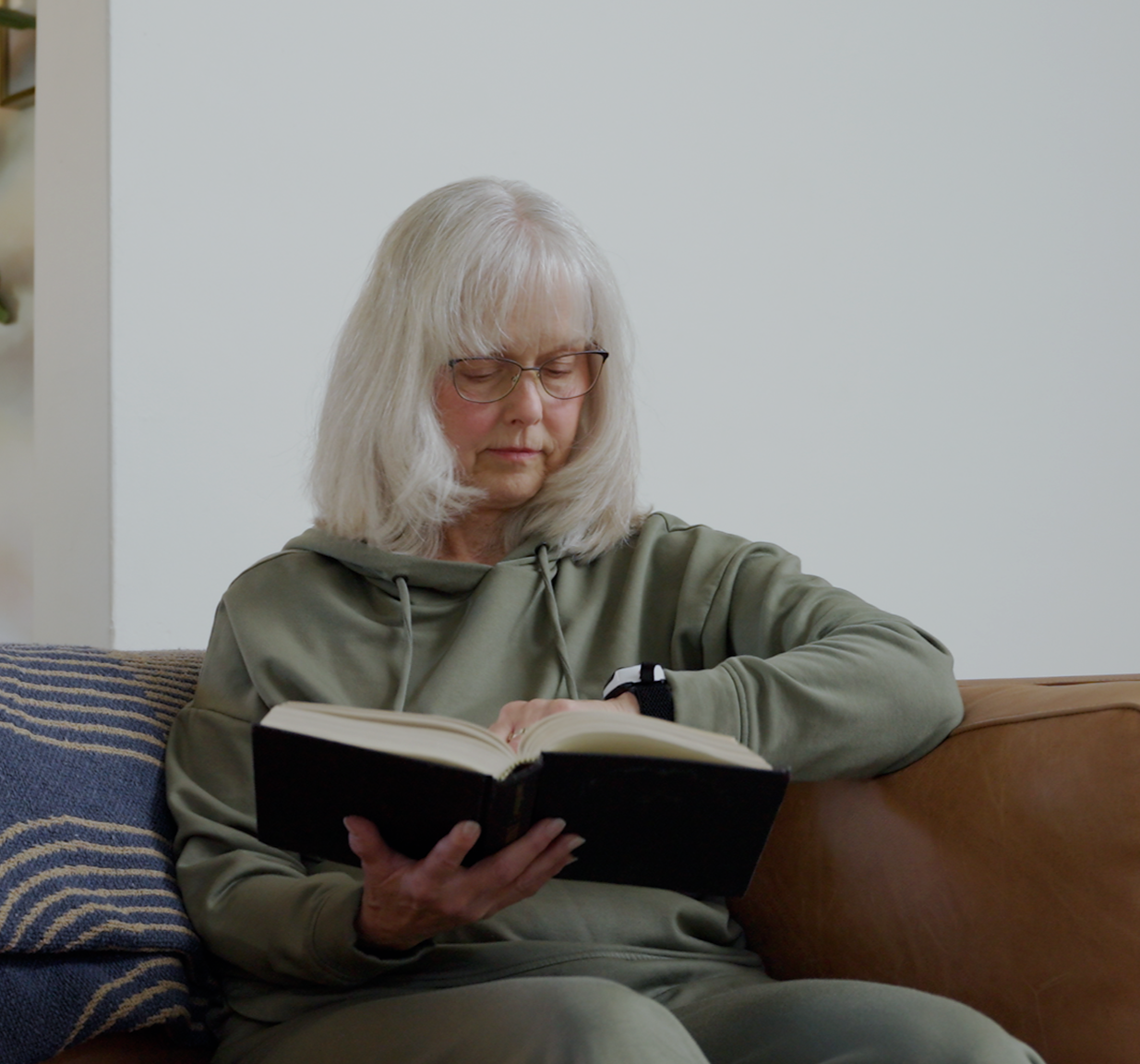 An older woman with white hair and glasses sits on a brown leather couch, reading an open black book. She is wearing an olive green hooded sweatshirt and matching sweatpants in a home setting.