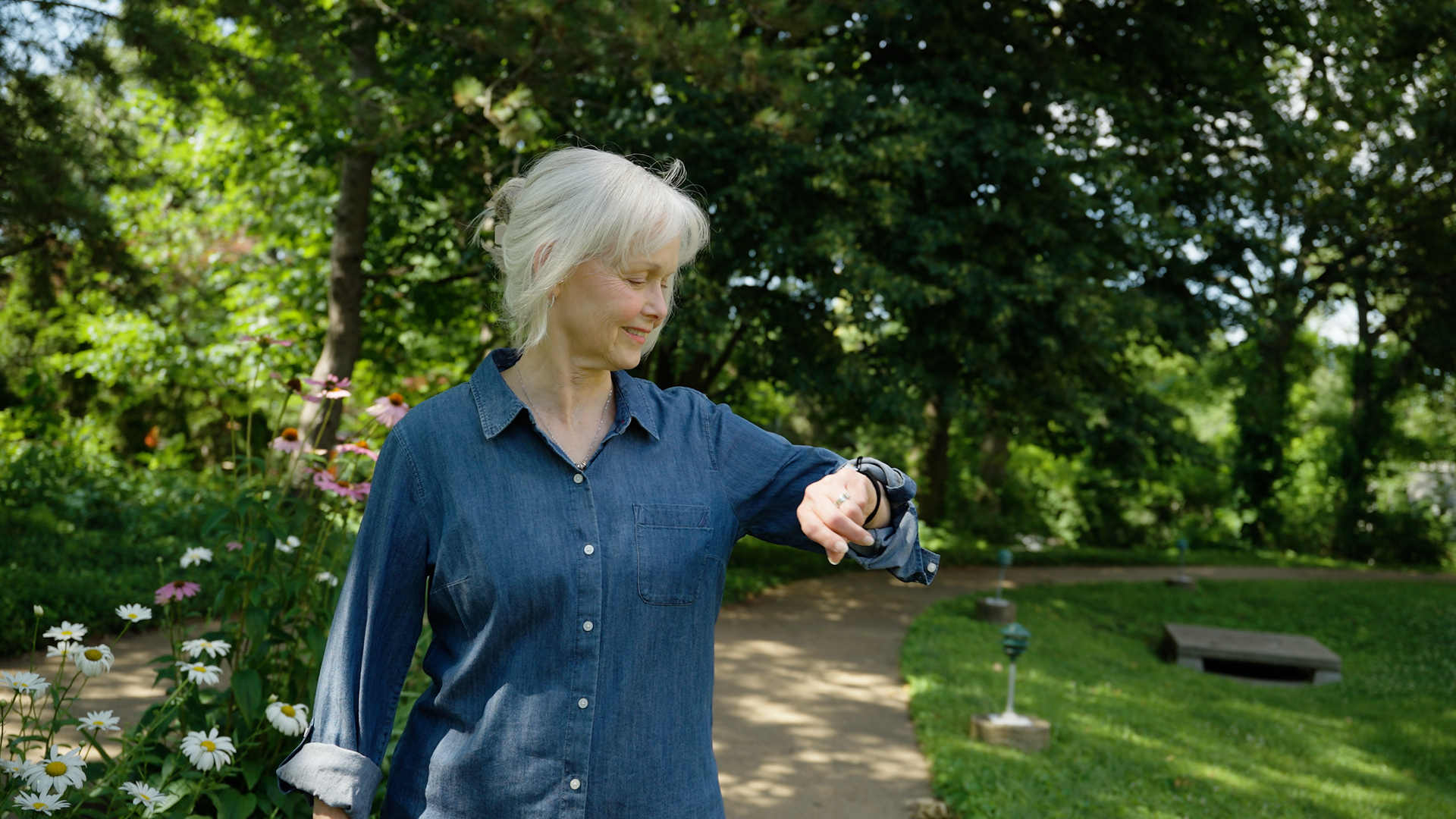A woman with short gray hair wearing a blue denim shirt walks in a park while looking at her SilverKin Tempo