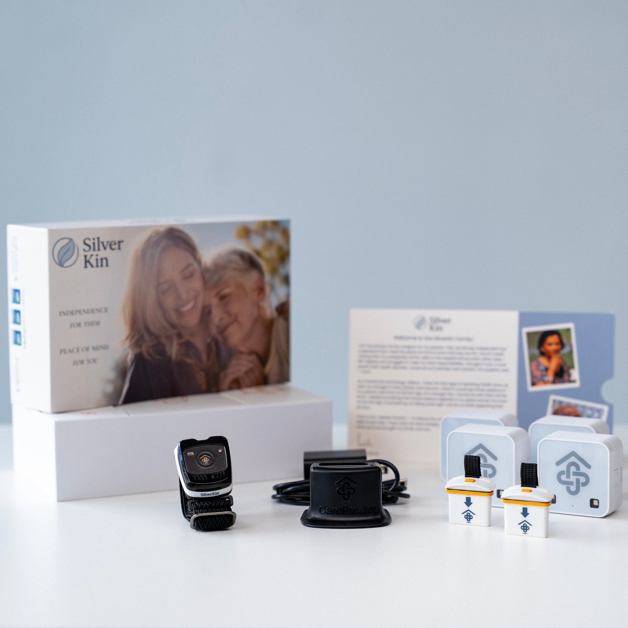 Silver Kin smartwatch care kit displayed on a white surface against a light blue-gray background, including the retail box, watch, charging dock, cables, and small accessory units with branded cards.