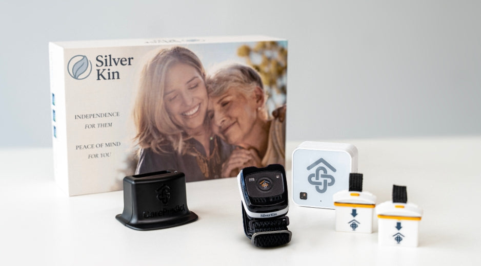 Silver Kin fall detection kit displayed on a white surface with its retail box. The kit includes a black wearable monitor, a black base, a white square receiver, and three small white battery-operated sensors with yellow bands and black tops.