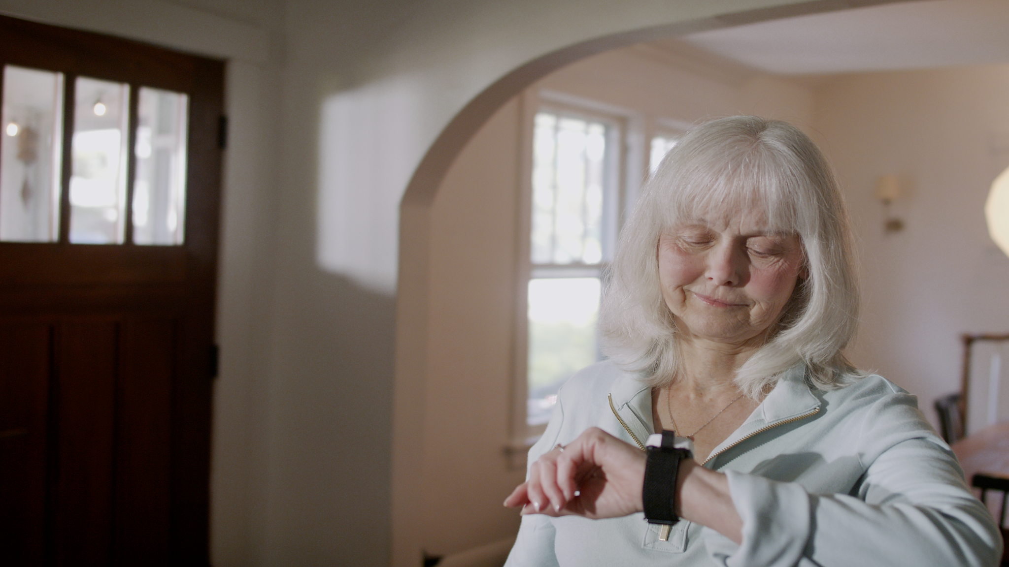 An older woman with white hair looks down at a black SilverKin Tempo her wrist, standing in a softly lit indoor room with cream walls and a wooden door in the background