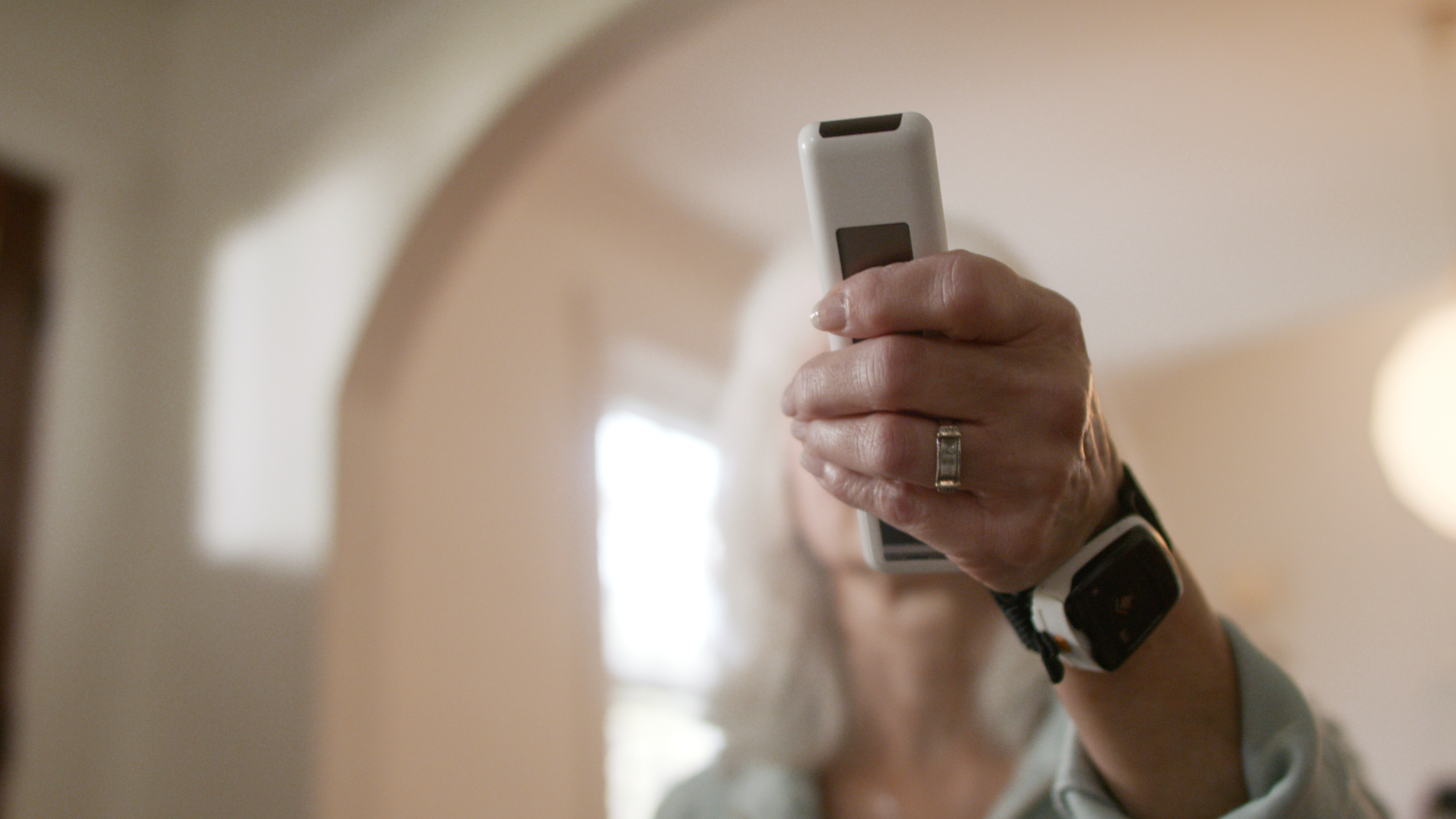 A close-up of a person's hand holding a white rectangular electronic device, with a blurred indoor background and an arched doorway behind them.